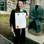 Aoife is wearing a graduation robe and mortar board and holding her degree certificate on front of her while standing outside NUI Galway.